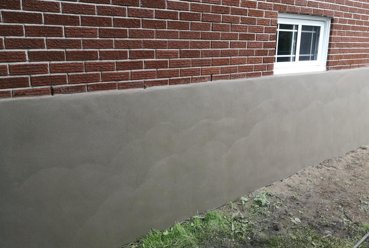 Brick building with newly applied, smooth gray stucco on the foundation wall, with a white-framed window.