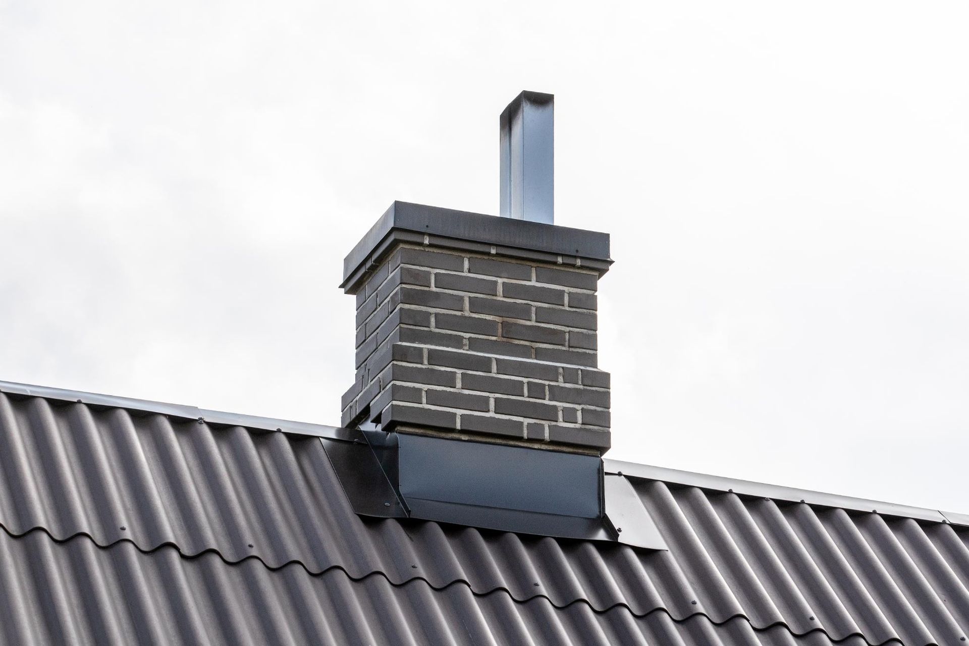 Brick chimney with a metal flue on a dark gray corrugated roof against a cloudy sky.