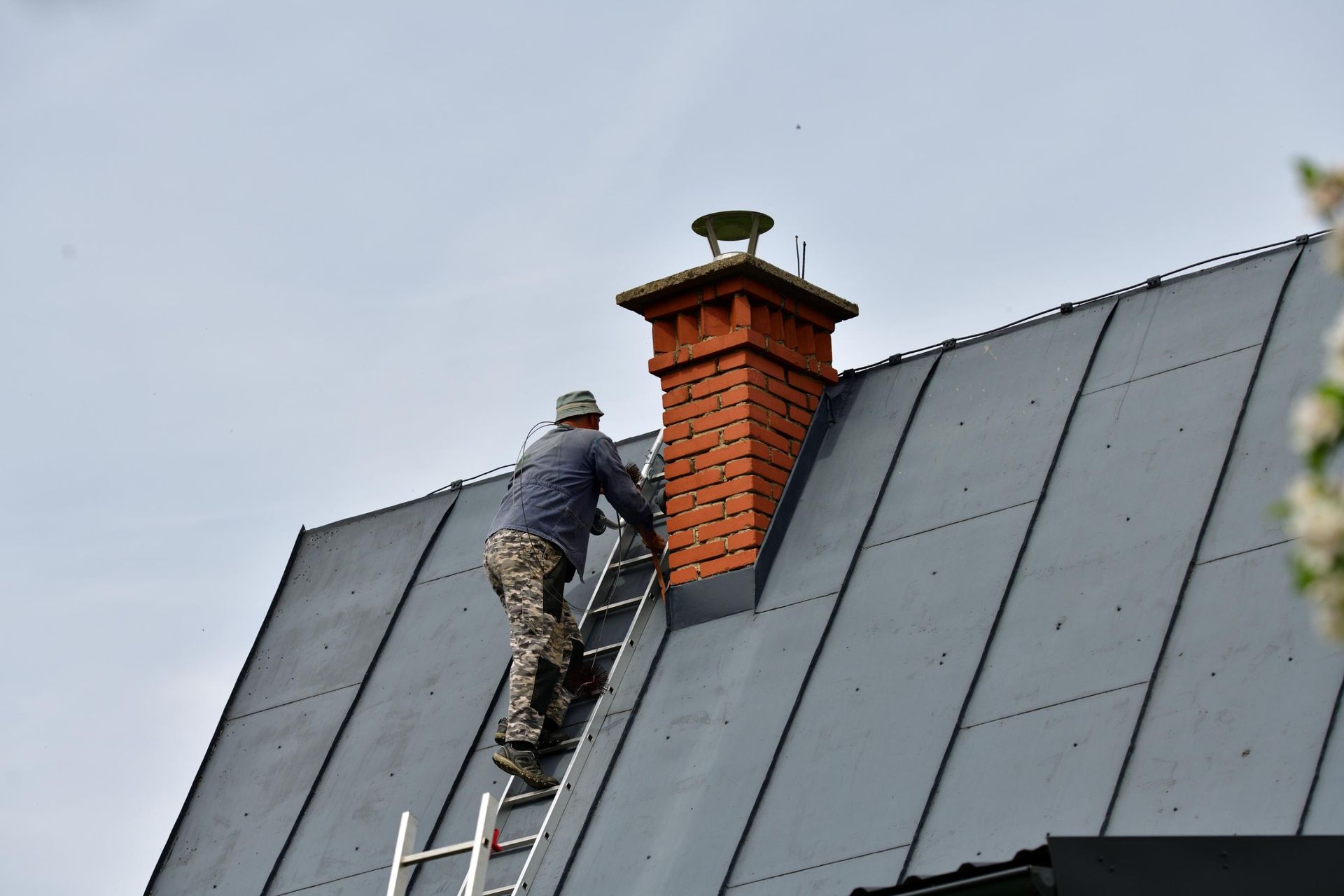 Man on a ladder, inspecting a red brick chimney on a gray metal roof under a cloudy sky.