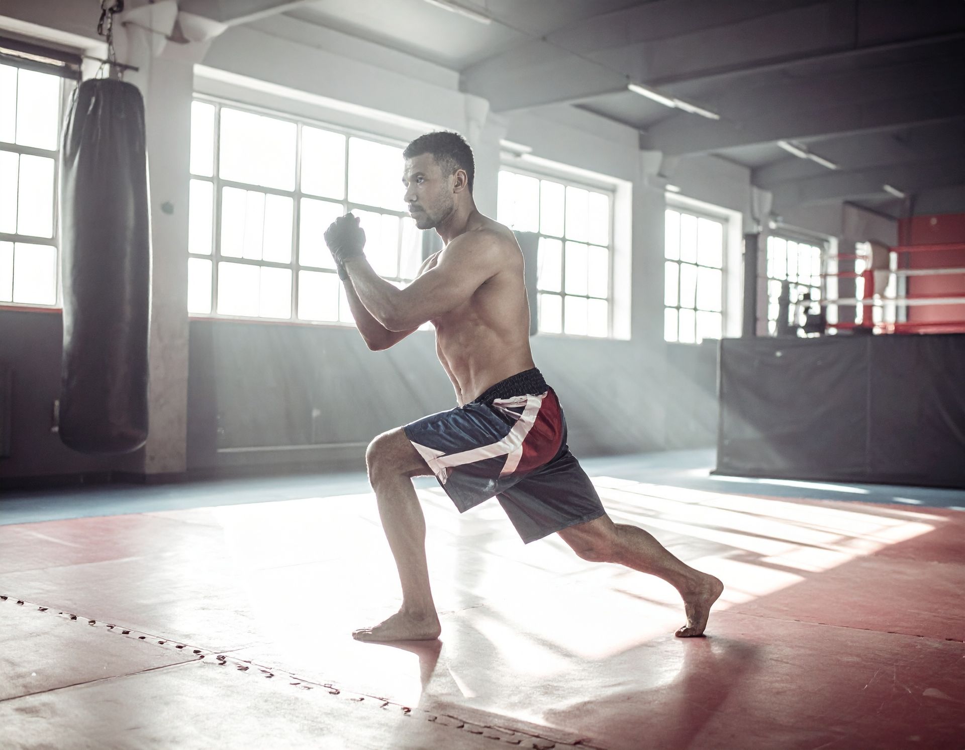 Bare-chested man in fighting stance, shadow boxing in a gym with punching bag and ring visible.