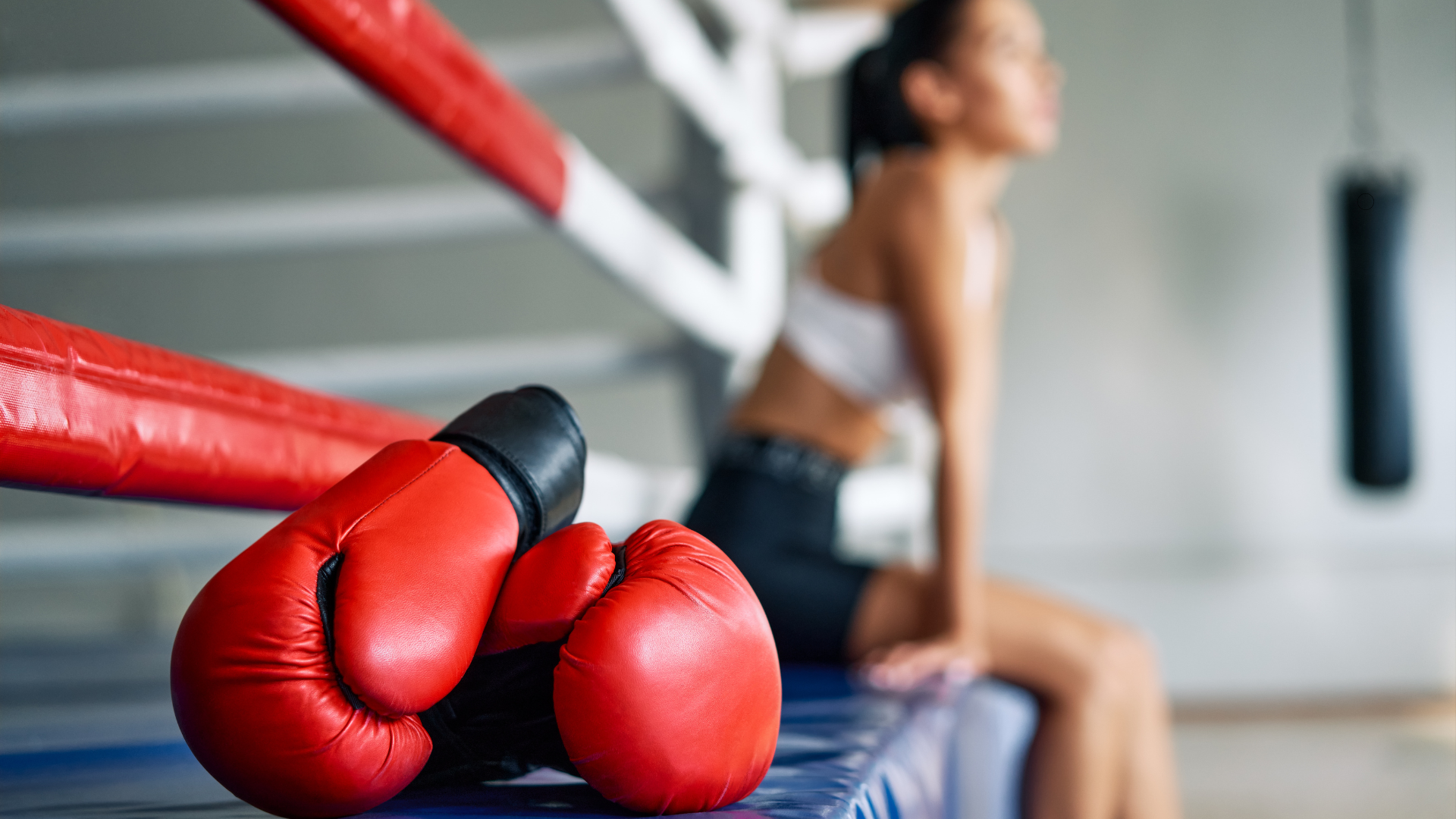 A woman is sitting on the edge of a boxing ring next to a pair of boxing gloves.