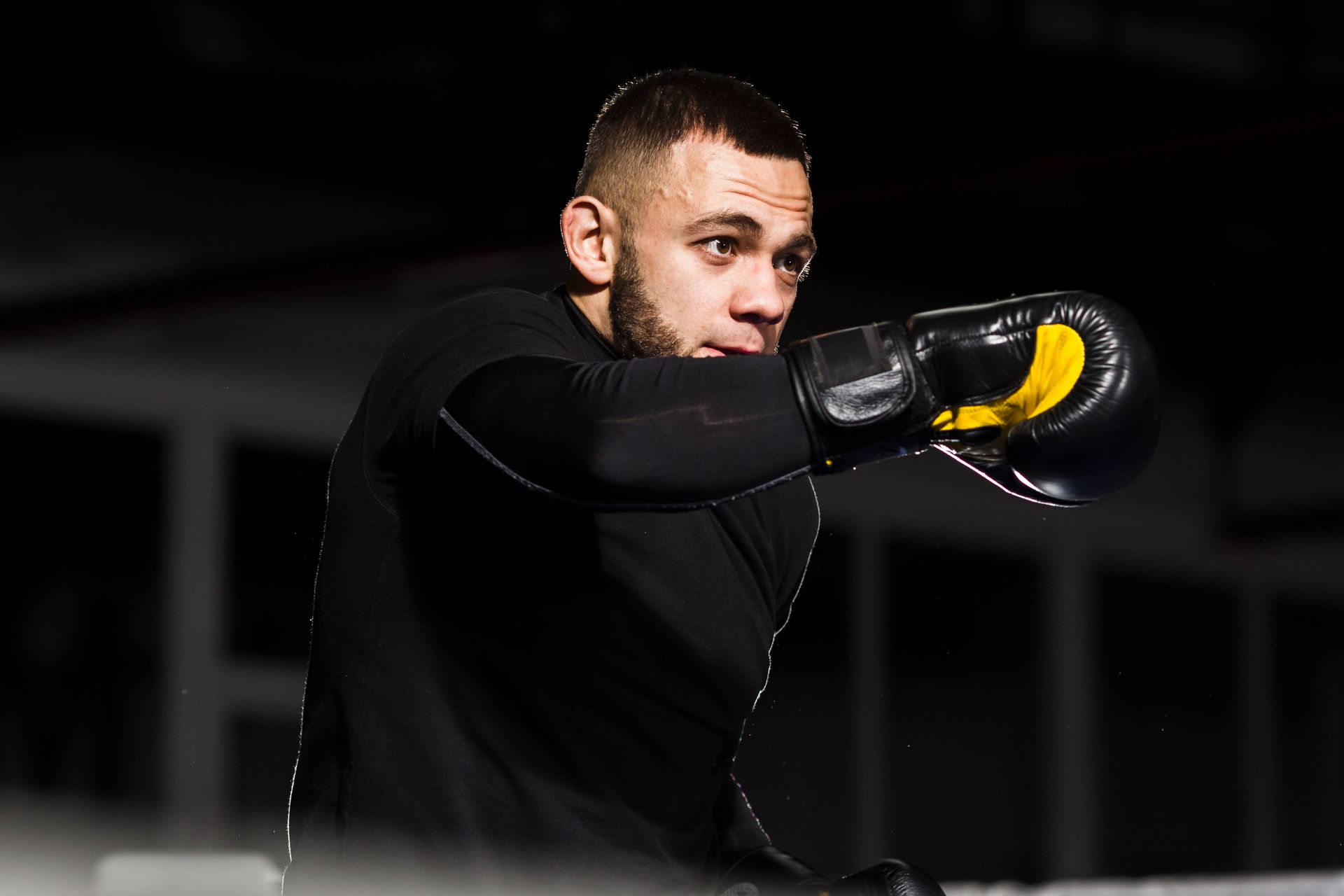 Boxer in black gloves throwing a punch, focused expression, dimly lit setting.