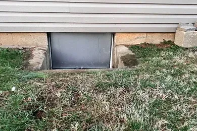 Basement window well under house siding with grass and dirt around it