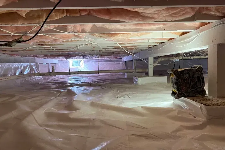 Plastic-covered crawlspace with exposed insulation, low beams, and a small window of daylight.
