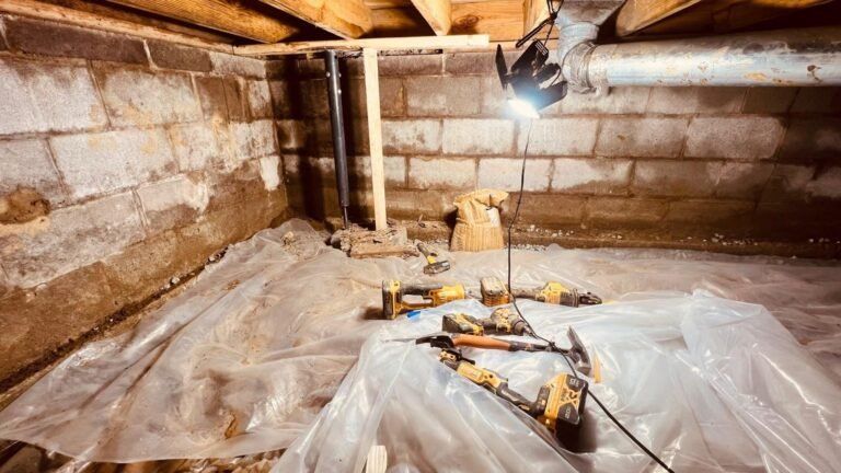 Basement under construction with plastic sheeting, tools, exposed walls, and a work light hanging overhead