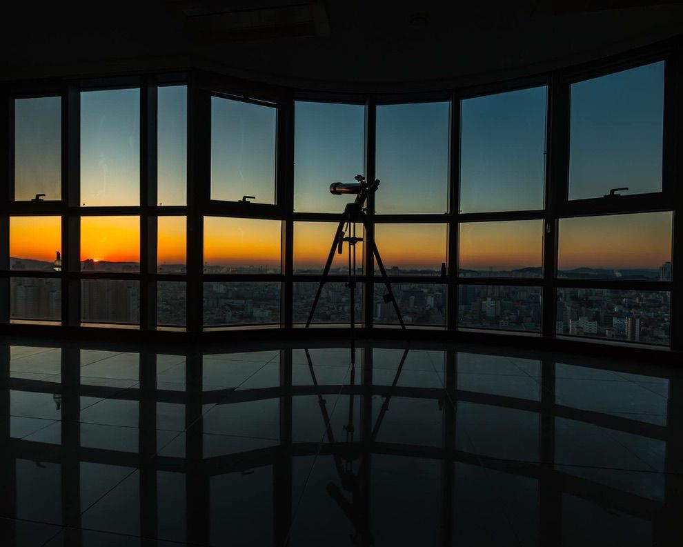 Tripod with a camera, silhouetted against a window with a sunset over a cityscape.