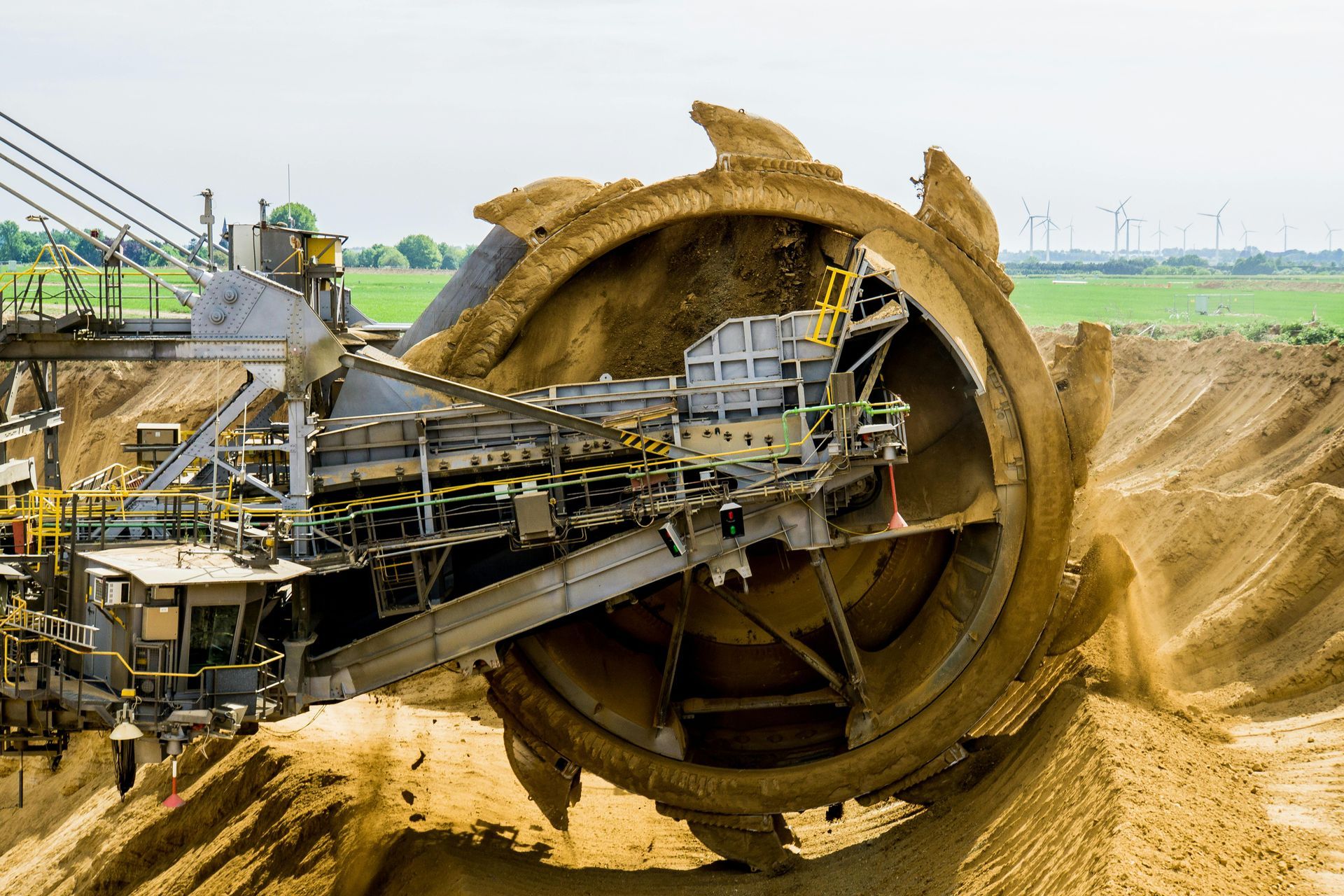 Large mechanical excavator digging into the earth, brown and tan colors with green field and windmills in the background.