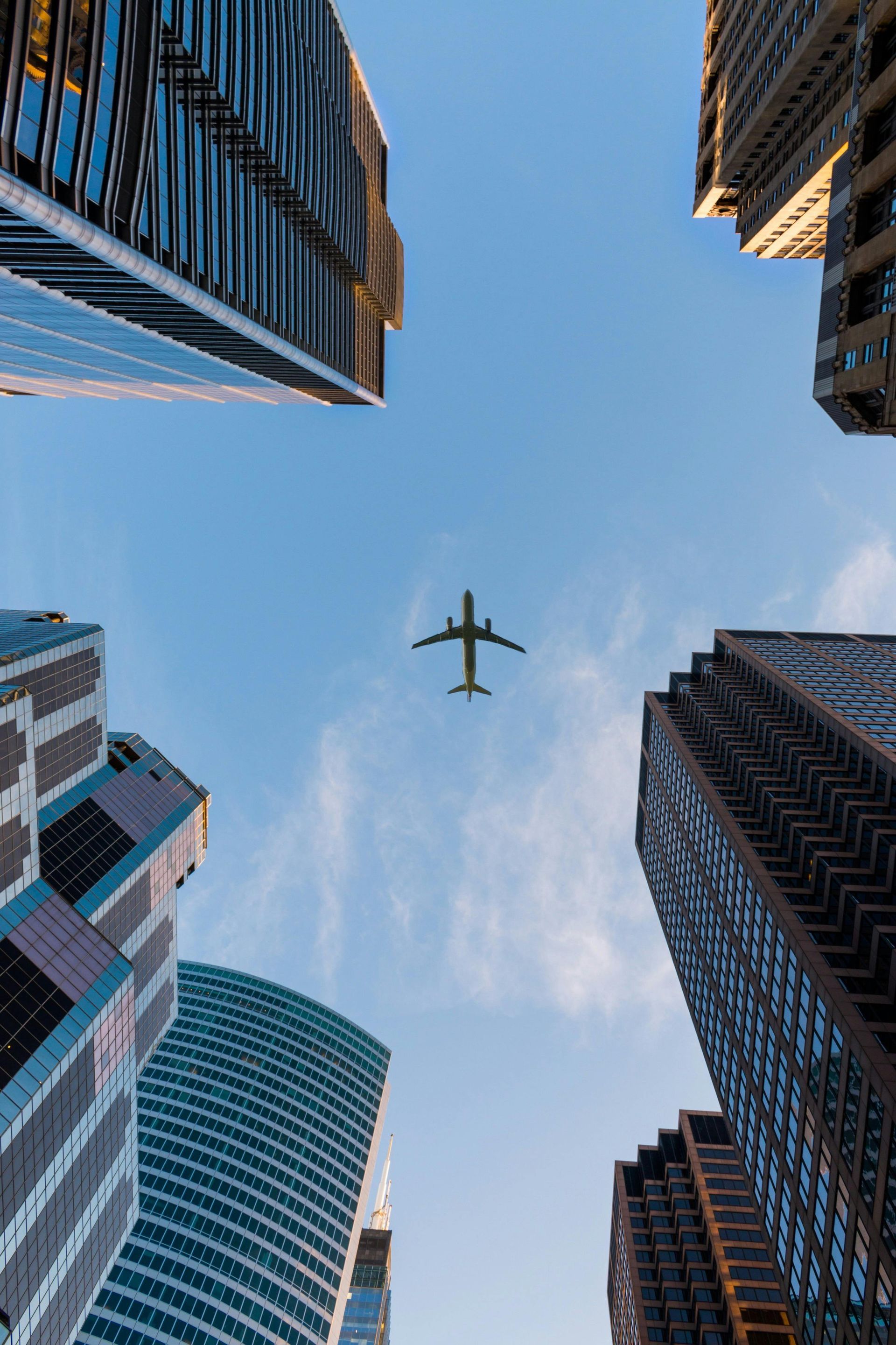 Skyscrapers framing the sky, an airplane flies overhead on a sunny day.
