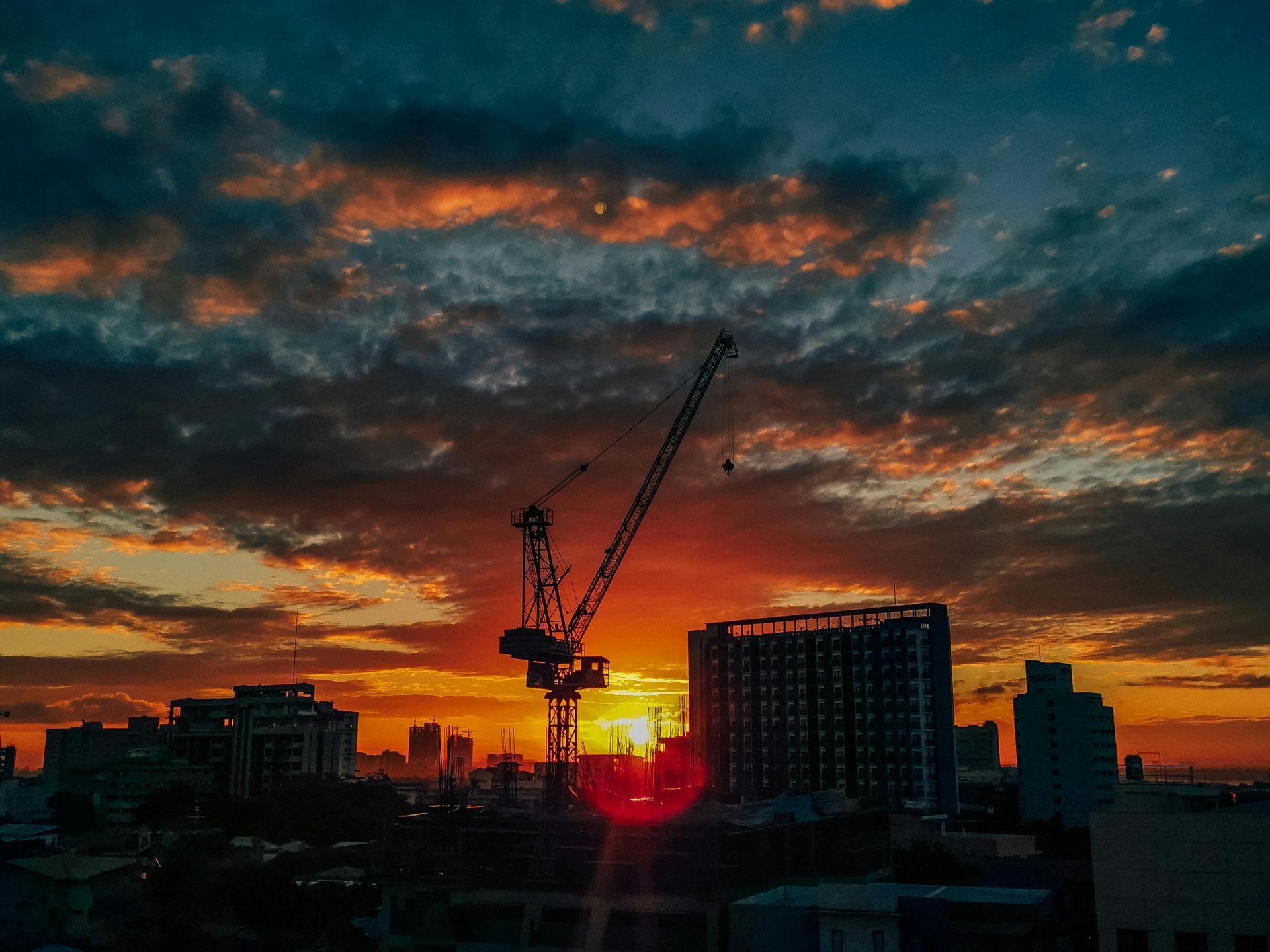 Sunset over a city skyline, silhouetted buildings and a crane against orange and blue clouds.