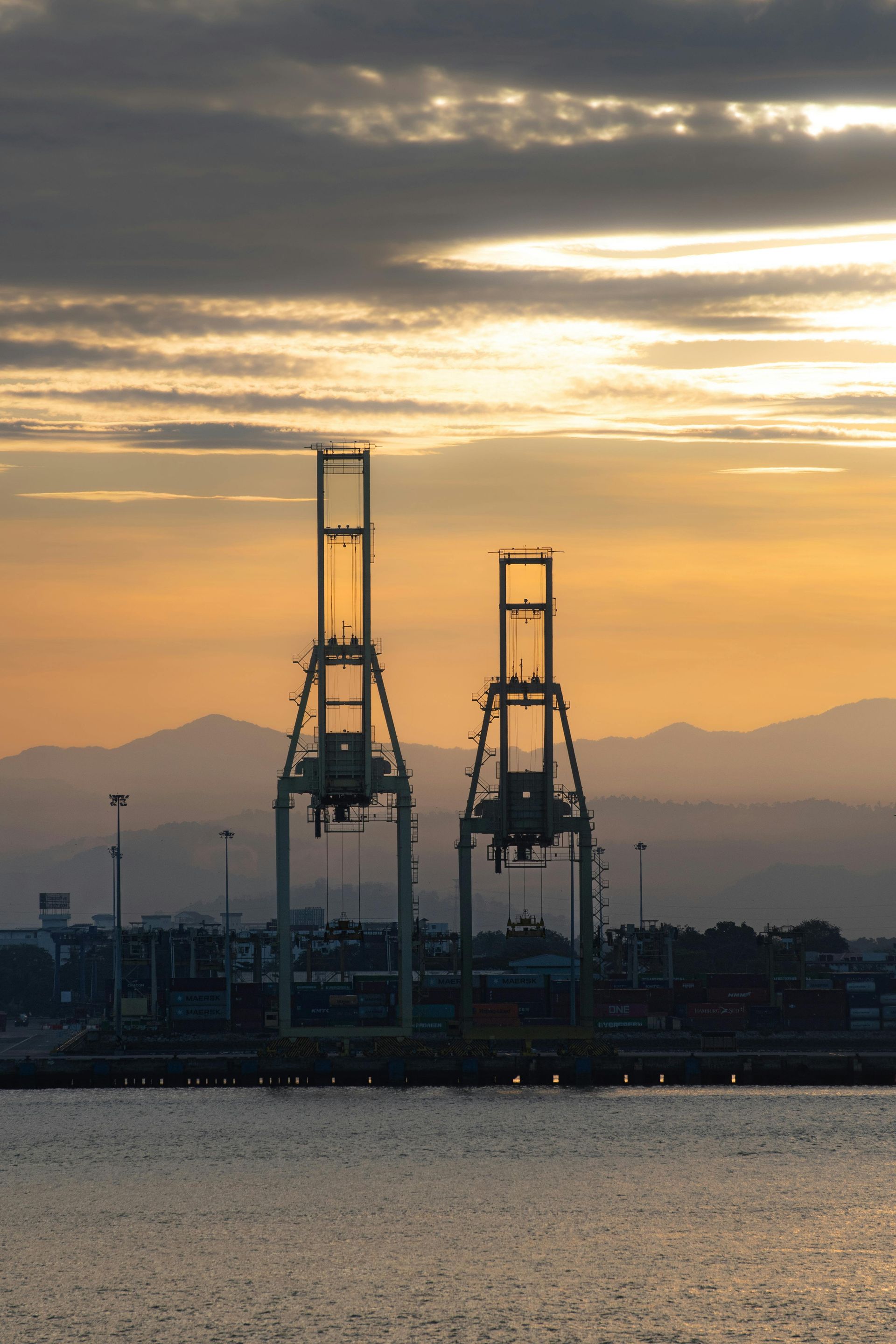 Two harbor cranes silhouetted against an orange sky with mountains in the background over water.