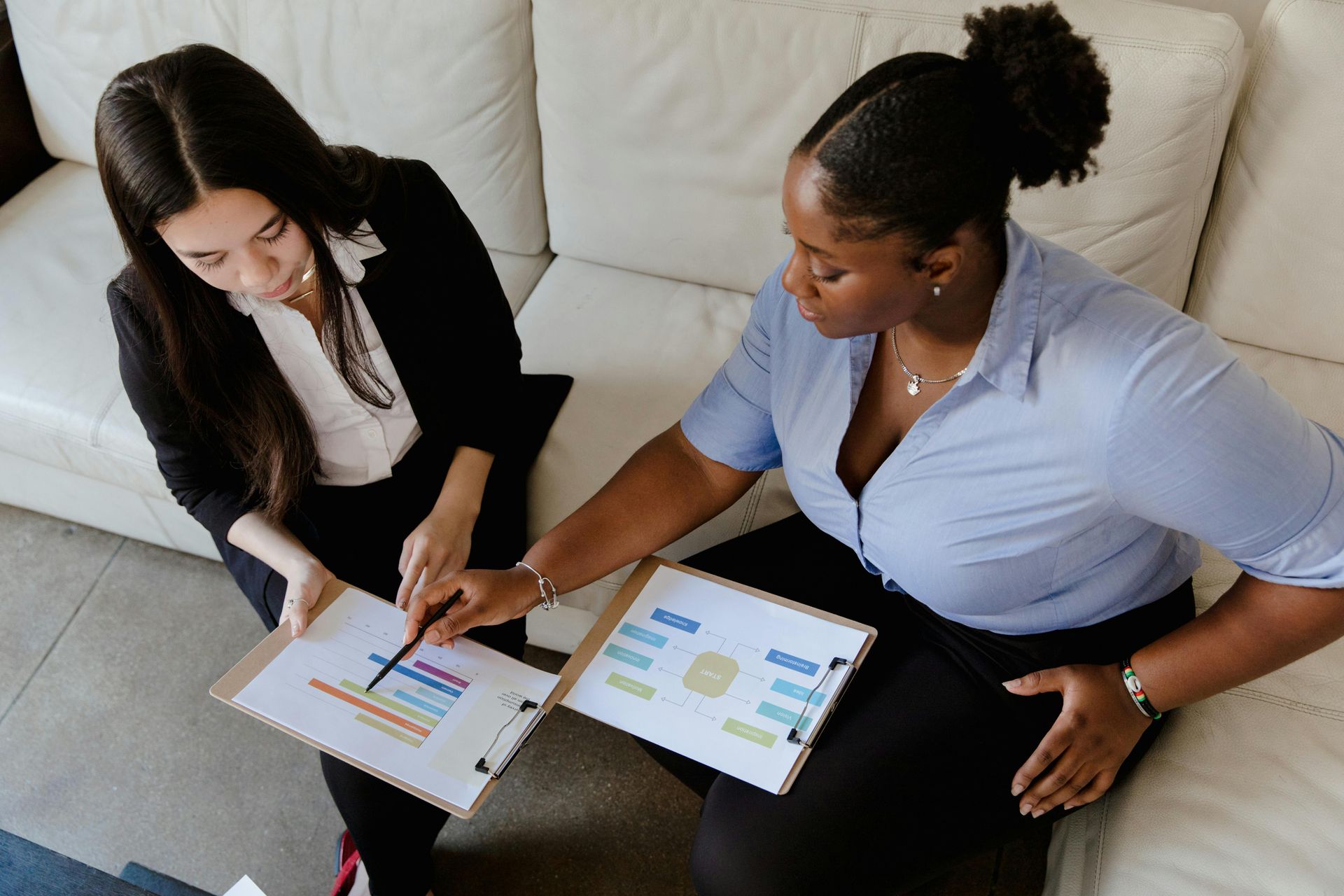 Two professionals sitting on a couch, reviewing and pointing at data charts on clipboards.
