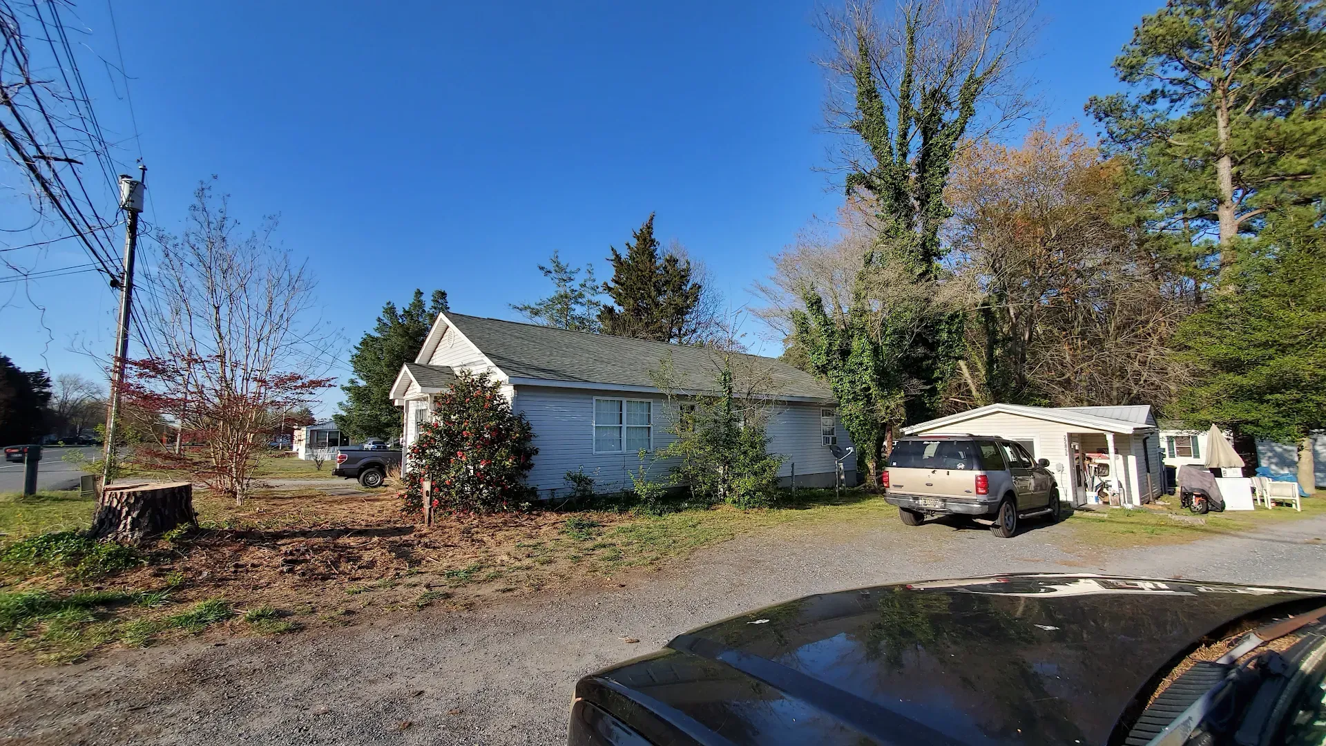 A small, weathered house with a dark roof and a vehicle parked in front, with trees in the background.