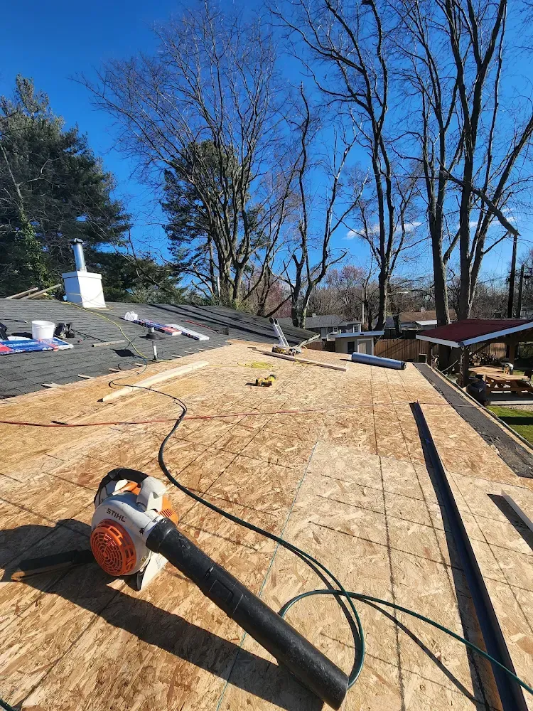 Roofing work in progress; new wooden sheeting laid, blower on roof under blue sky with trees.