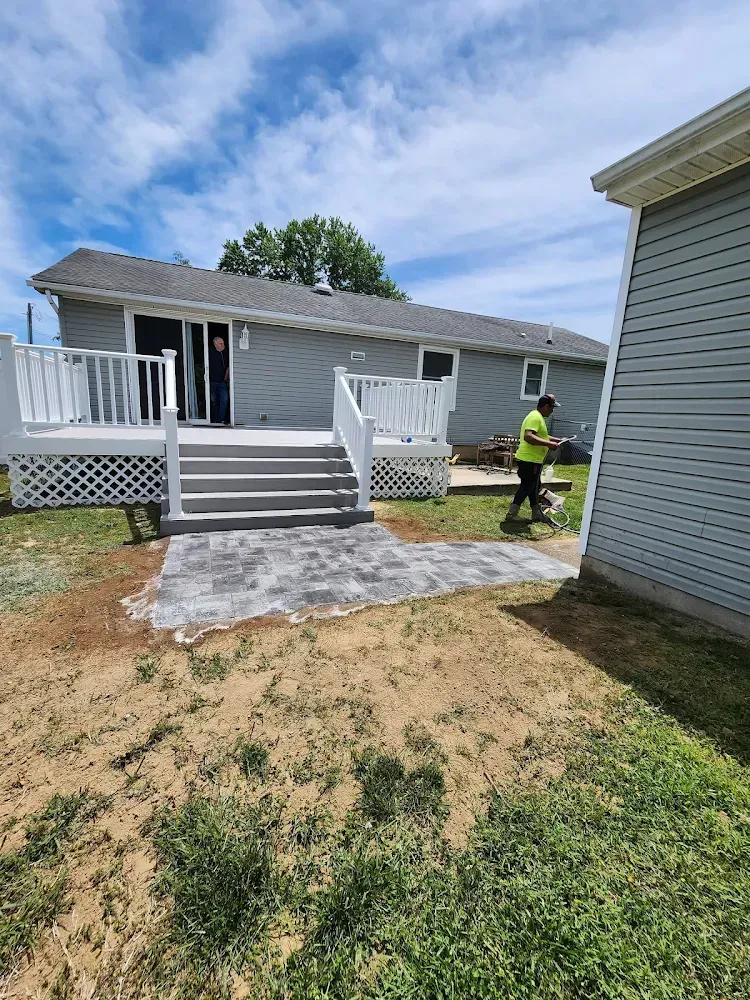 Backyard with gray house, deck, and patio. Person walks near a shed, blue sky with clouds.