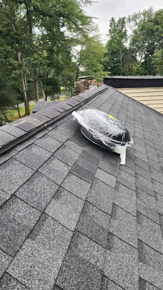 Black roof vent on asphalt shingle roof, wrapped in plastic. Trees in the background.