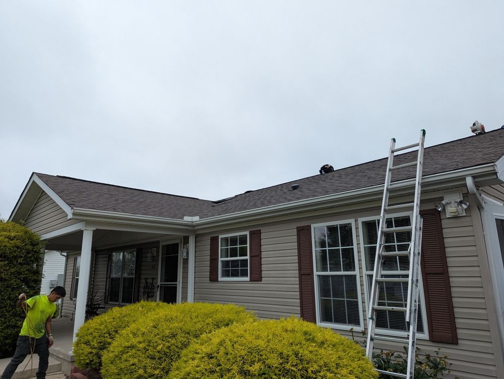 Man on roof, ladder at side of house. Overcast sky, brown roof and shutters, beige siding.