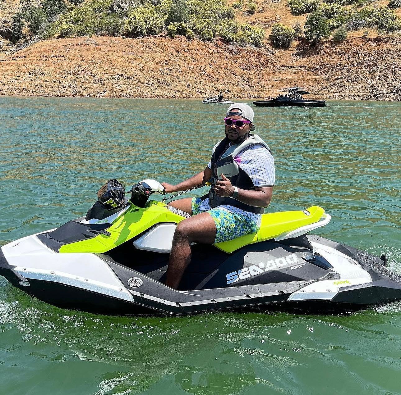A person wearing a white hat, sunglasses, and patterned shorts sits on a yellow and grey Sea-Doo jet ski on a lake.