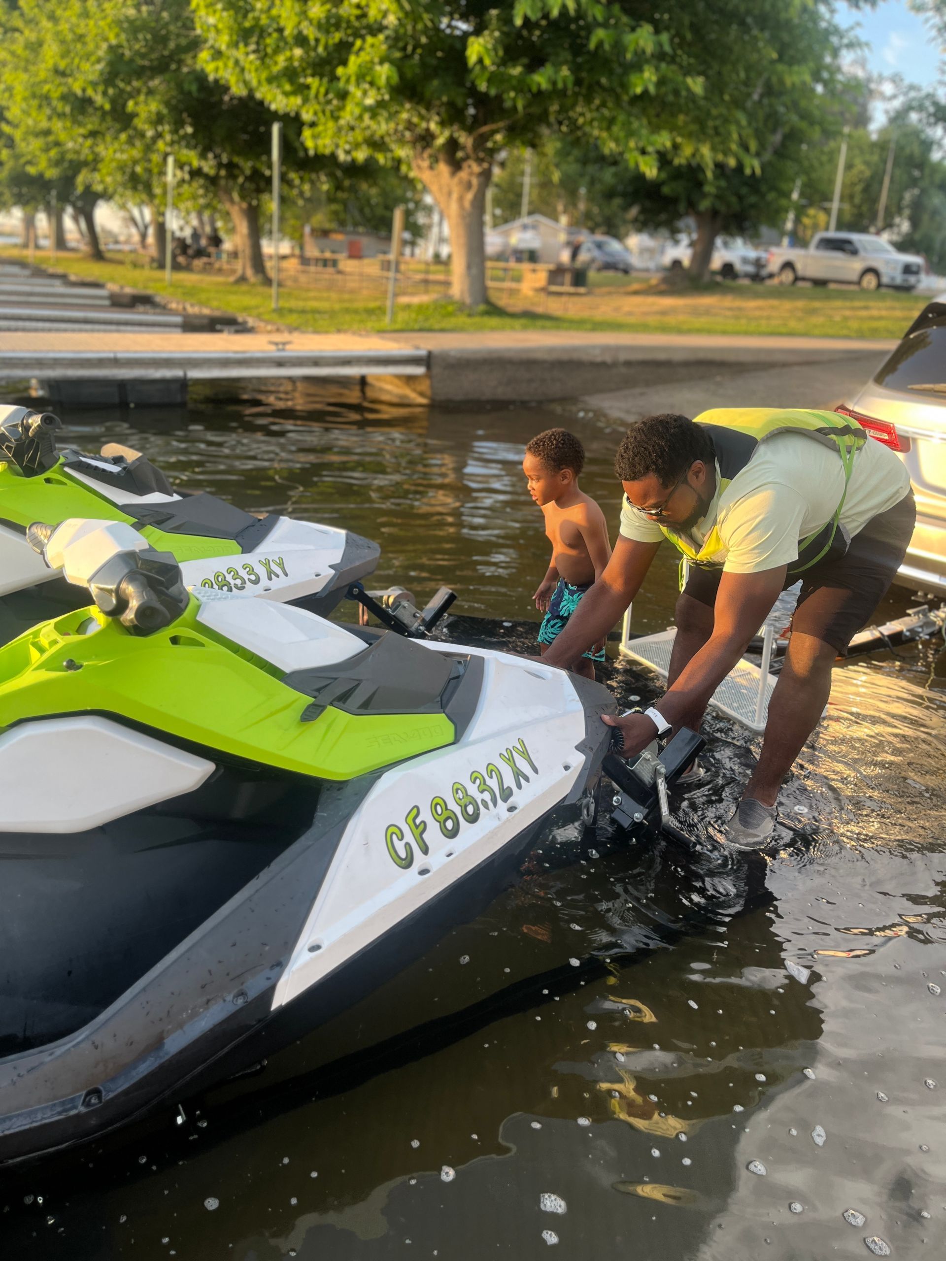 A person in a yellow life vest assists a child in launching a lime green and white jet ski at a boat ramp.