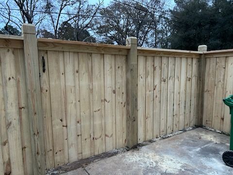 Wooden fence corner on concrete patio, surrounding a green trash can, overcast sky.