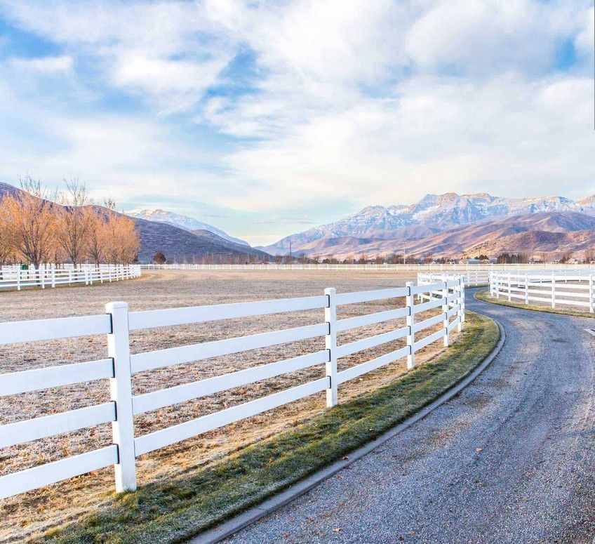 A white fence surrounds a dirt road with mountains in the background.