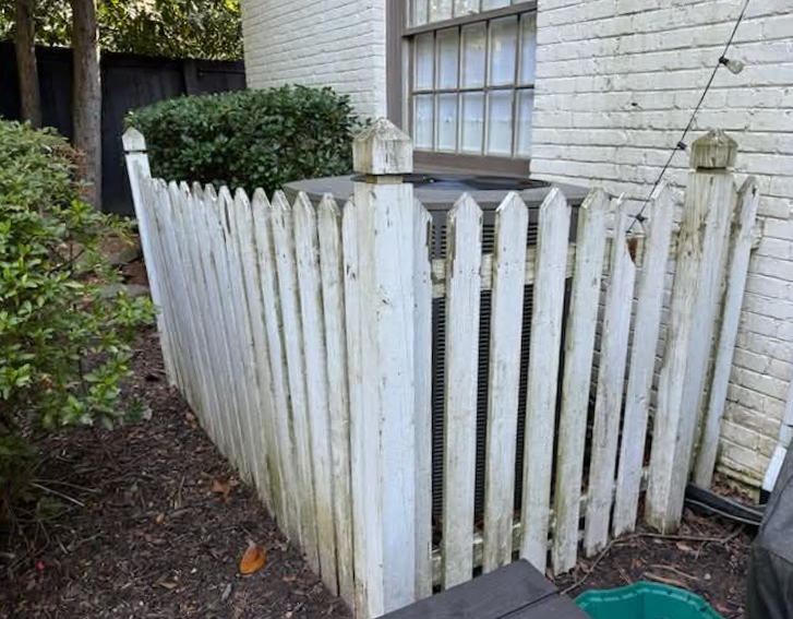 White picket fence encloses an air conditioning unit next to a brick building.