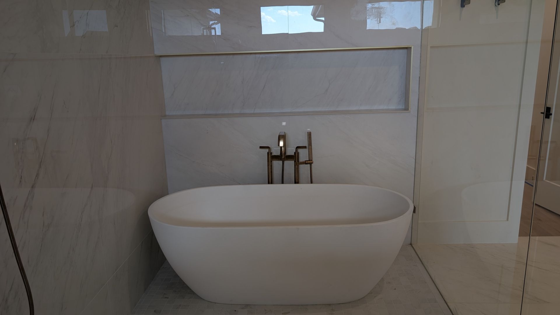Modern white bathtub with brushed gold faucet, set against white marble-tiled wall with recessed shelf.