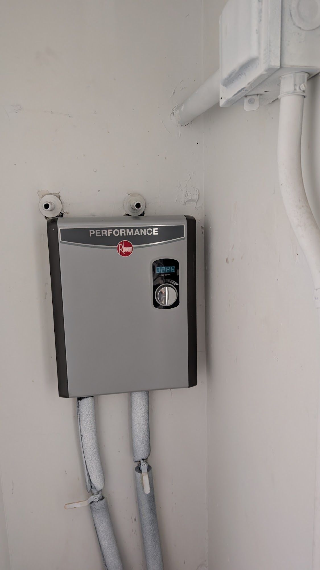 Gray electric water heater mounted on a white wall in a corner. Water pipes below.