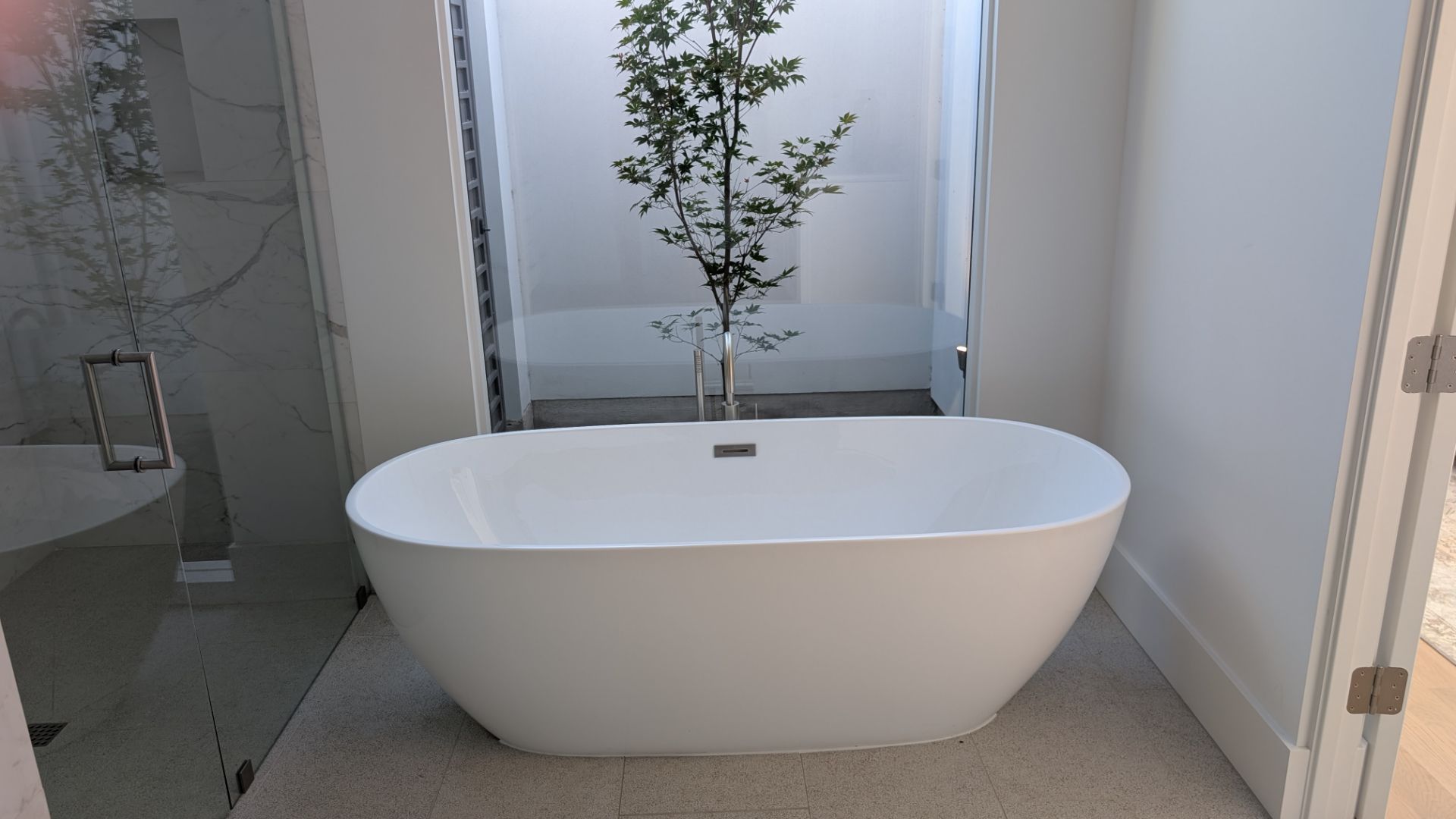 White oval freestanding bathtub in a bright bathroom with a window and a small tree.