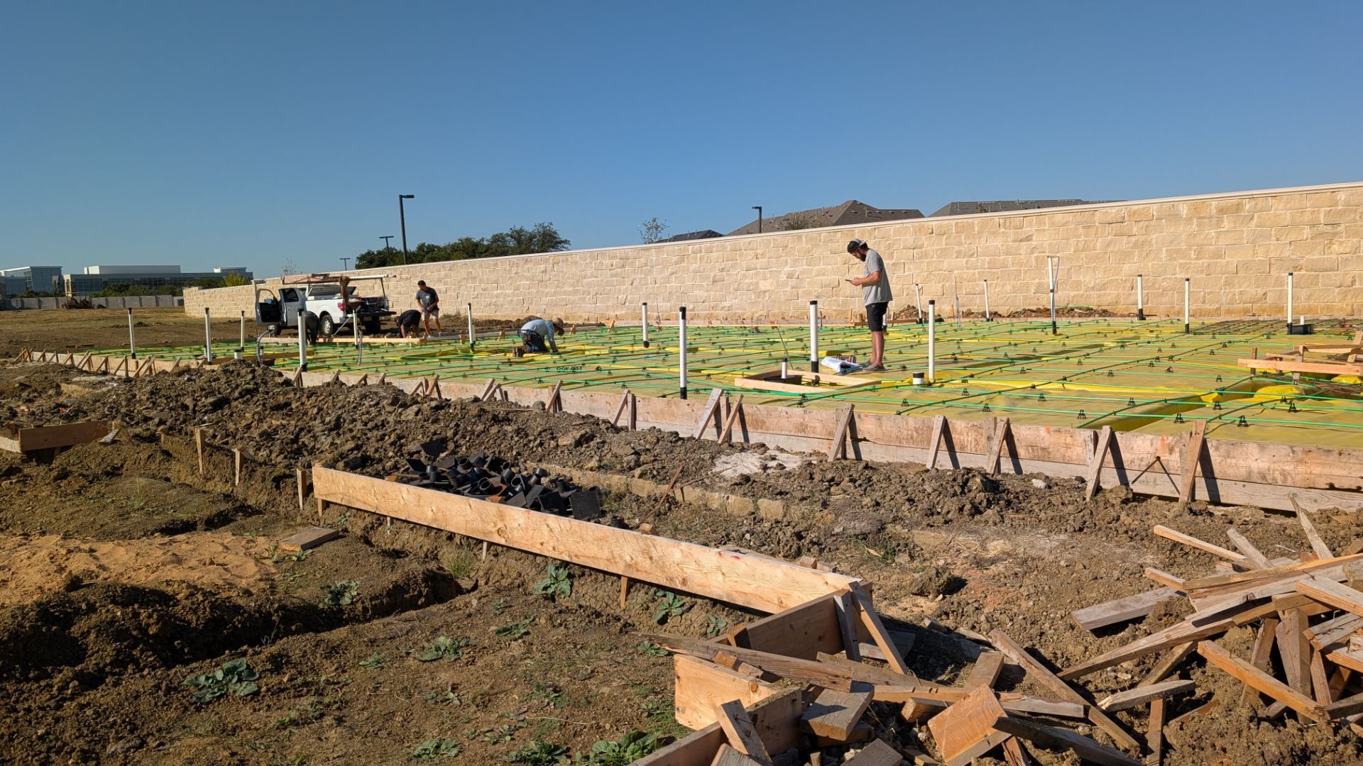 Construction site: Workers building forms, surrounded by soil and materials. A wall and blue sky are in the background.