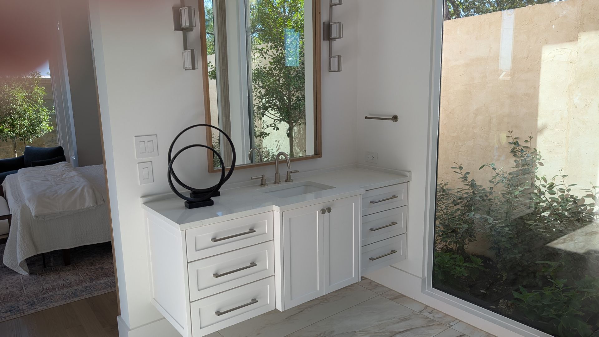White bathroom with vanity, mirror, and a view of greenery.