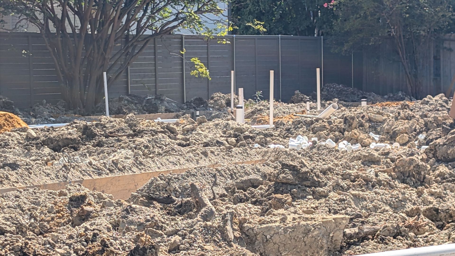 Excavated construction site with exposed pipes and a wooden fence backdrop.