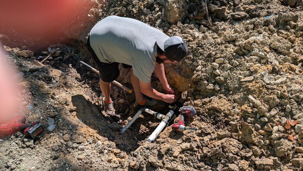 Person working on pipes in a dirt hole outdoors, wearing a gray shirt and hat, sunny.