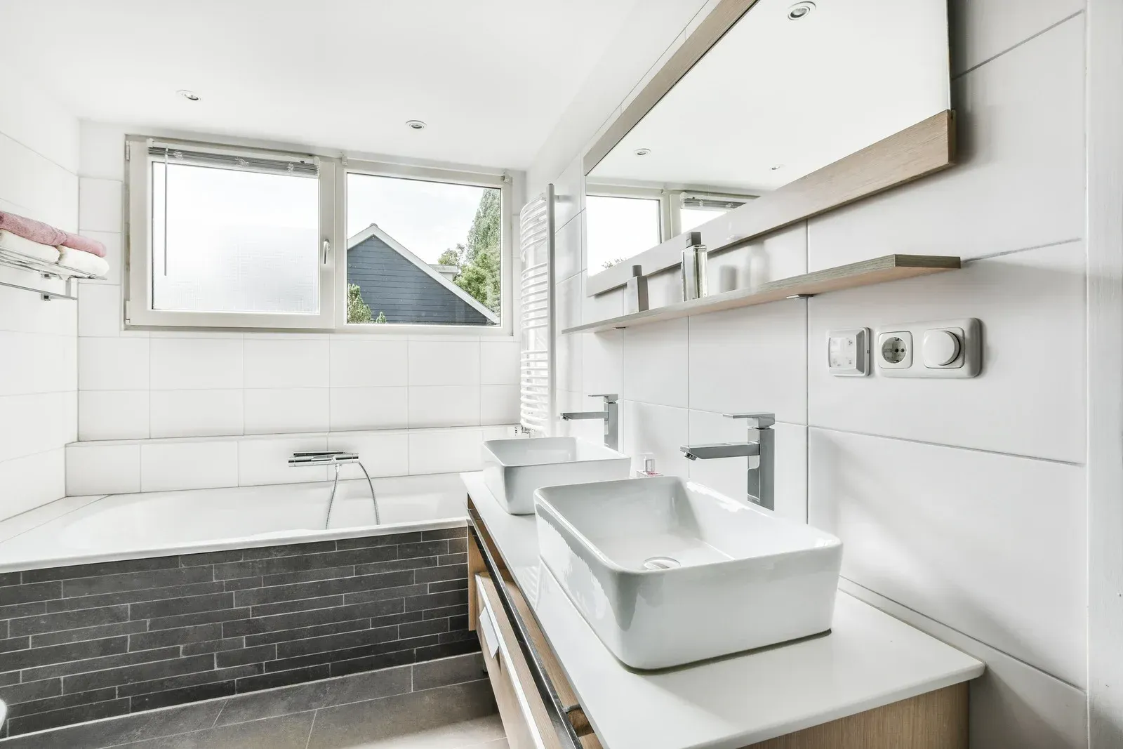 Modern bathroom with white sinks, tub, and tile. A large mirror and window provide natural light.