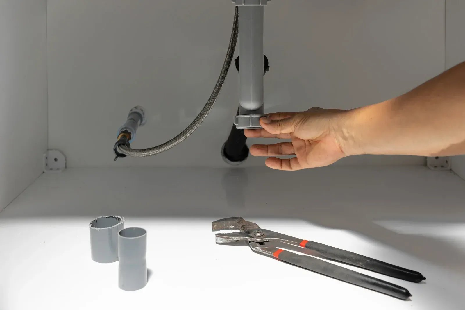 Person's hand adjusting a sink drain pipe inside a white cabinet. Pliers and pipes are in the cabinet.