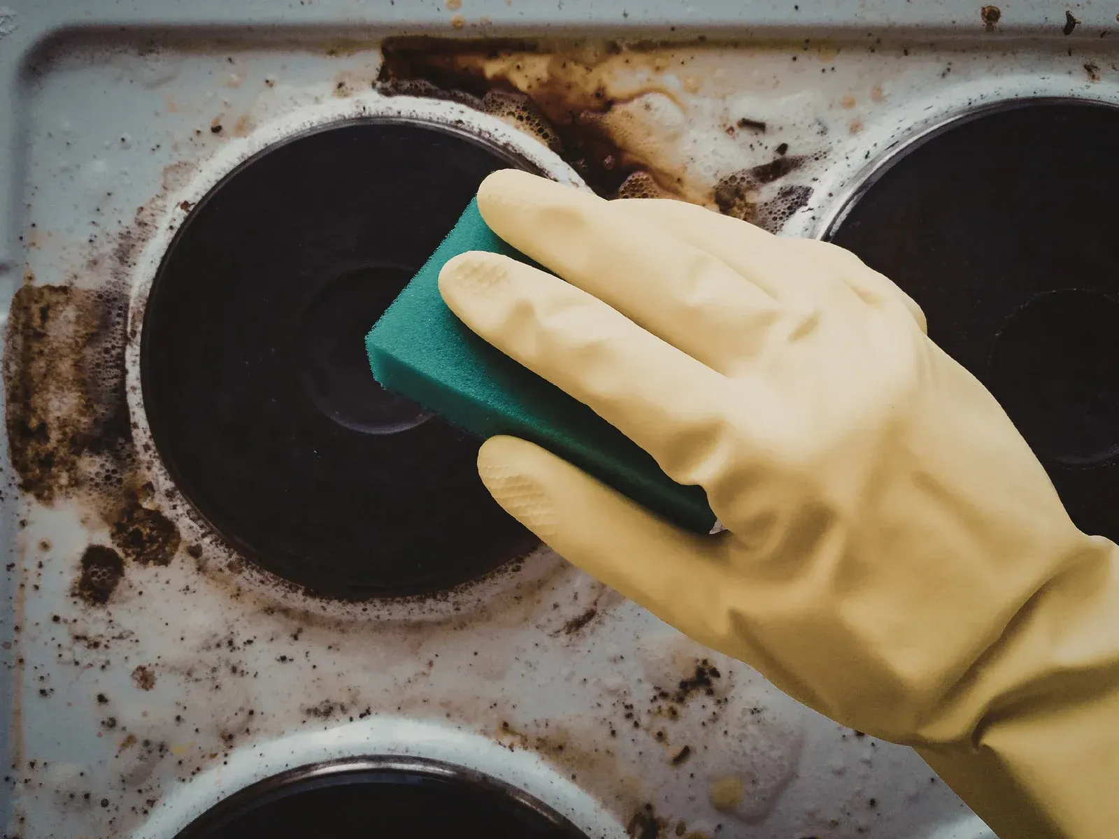 Gloved hand with sponge cleans a dirty stovetop with burnt food residue around the burners.