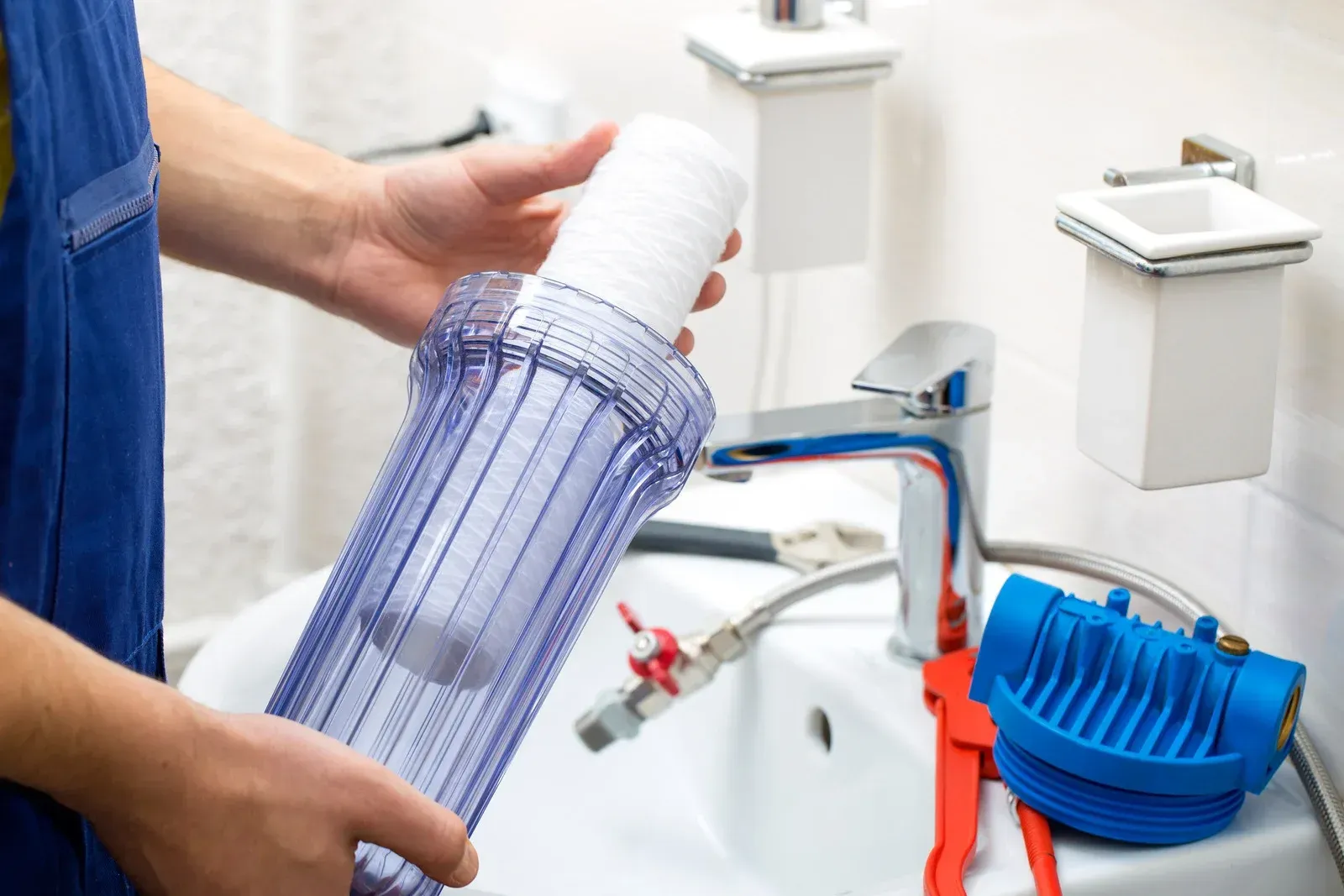 Plumber holding water filter, sink in background, blue overalls, water faucet, and wrench visible.