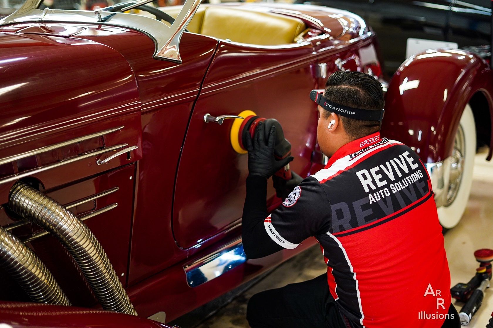 A man in a red and black shirt polishes a classic maroon car door with a buffer.