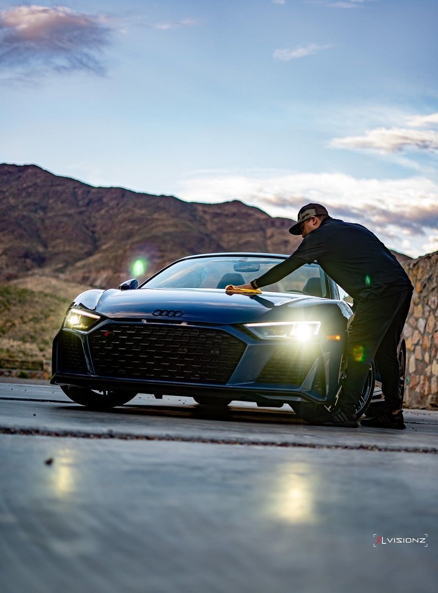 Man leaning on a blue Audi R8 sports car on a road in front of mountains.