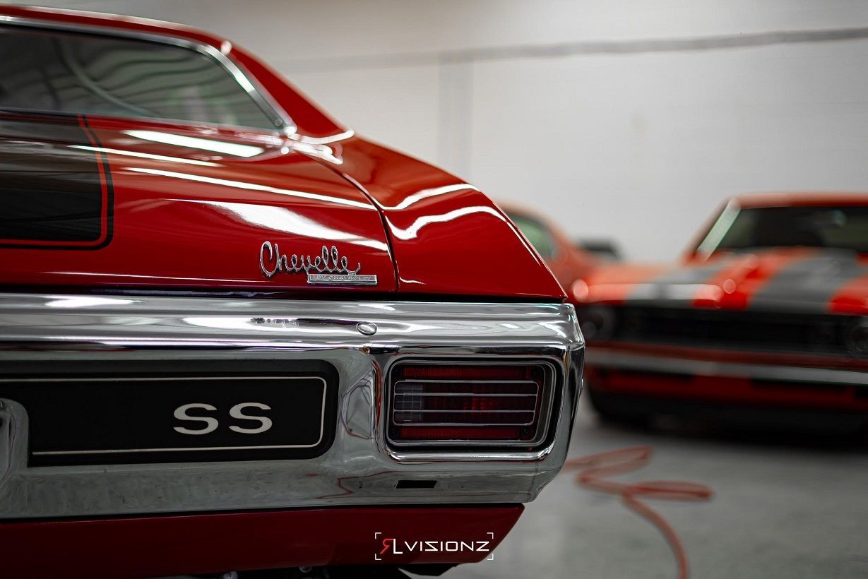 Red Chevrolet Chevelle SS muscle car. Rear view, black stripe, chrome detail, another car blurred in background.