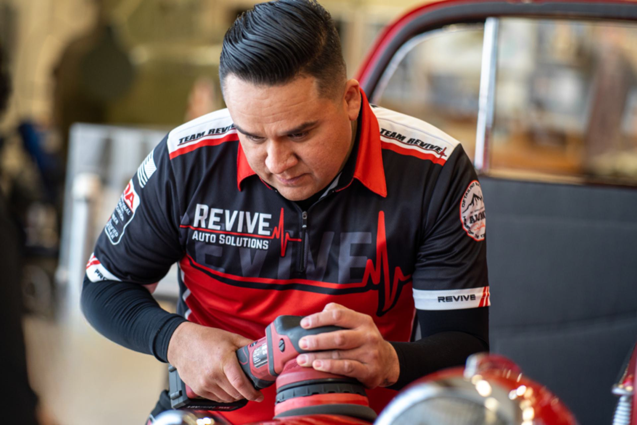 Man polishes a car with a buffer, wearing a Revive Auto Detailing shirt, in a workshop setting.