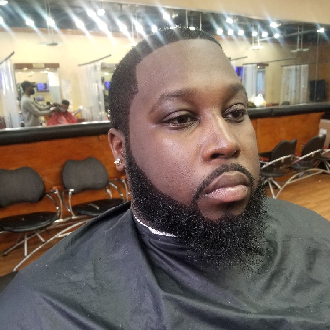 Man with a groomed beard and haircut, sitting in a barber shop, looking at the camera.