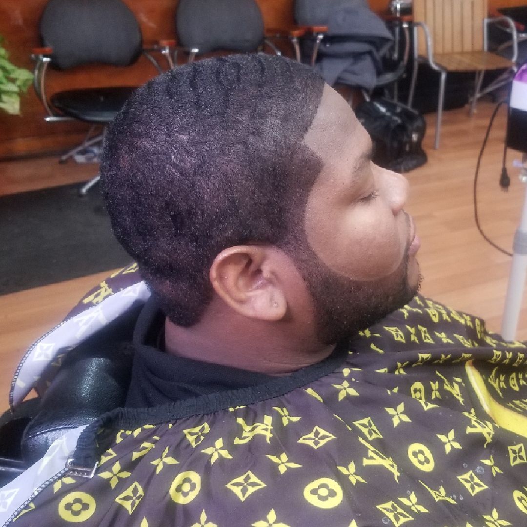 Man with a fresh haircut and beard trim, in a barbershop. He wears a cape with a gold pattern.