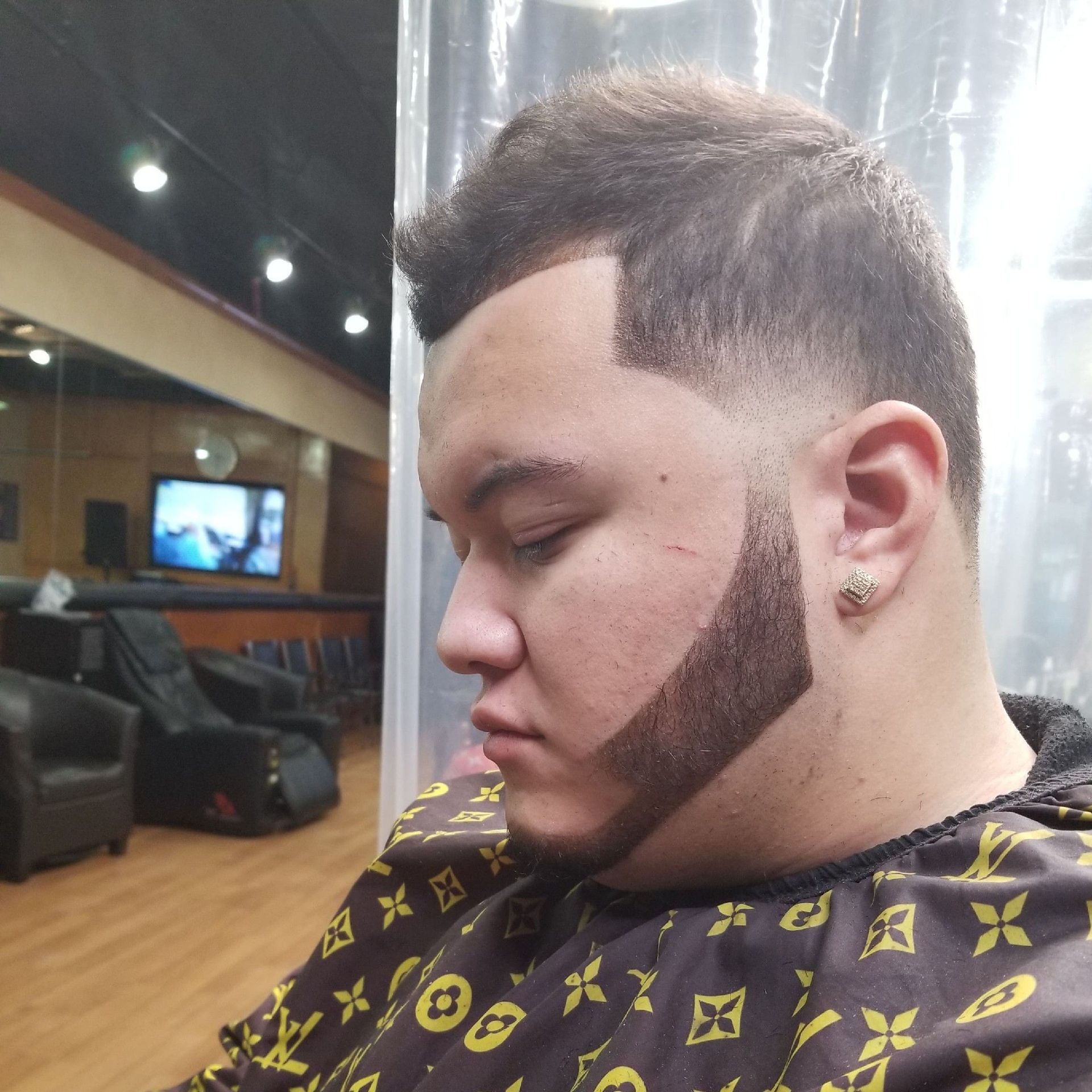 Man with a trimmed beard and faded haircut in a barber shop setting, wearing a patterned cape.