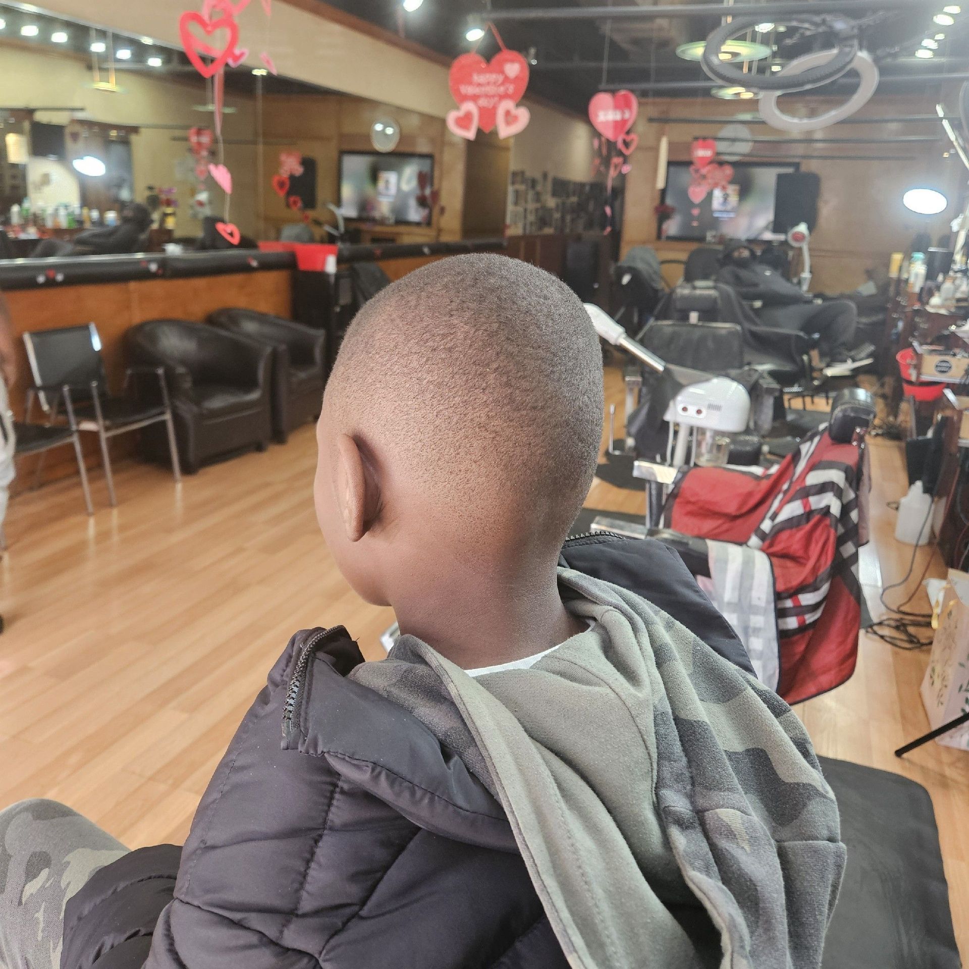 Child with a short haircut in a barbershop, facing away from the camera. The setting is indoors, with chairs and mirrors.