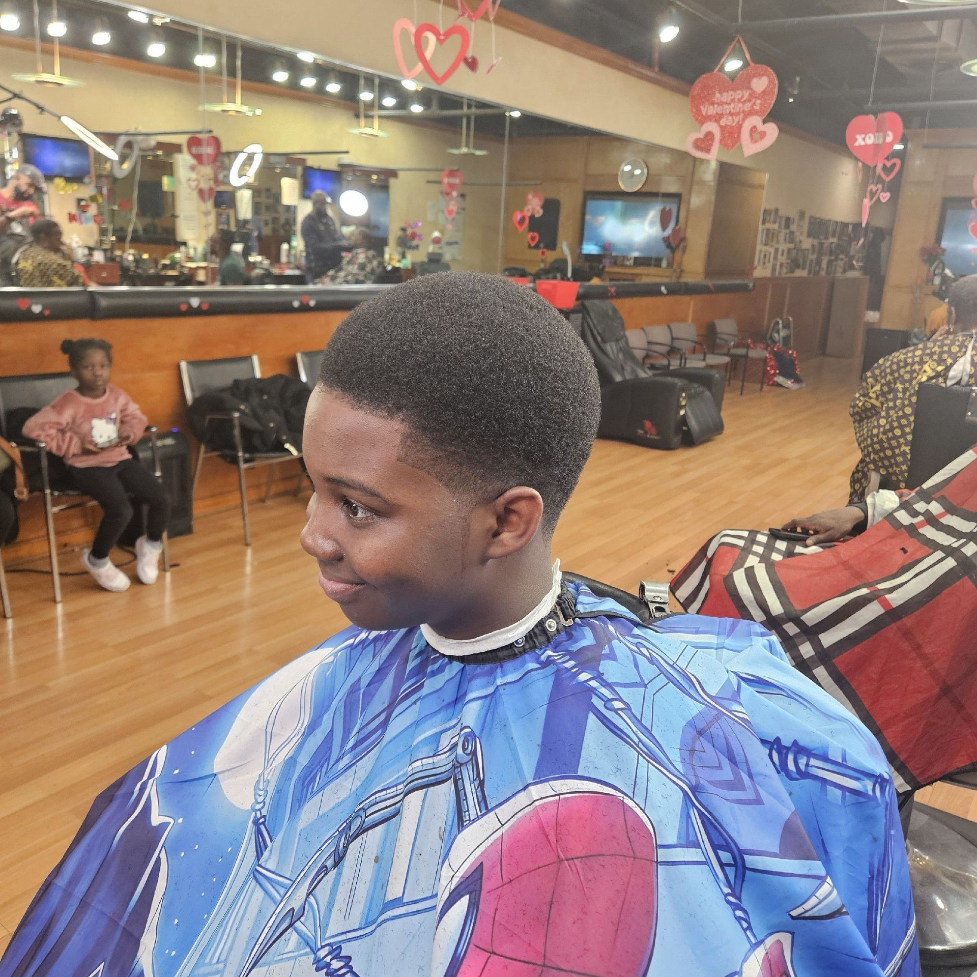 Boy with a short haircut in a barber shop, wearing a Spider-Man cape, smiling.