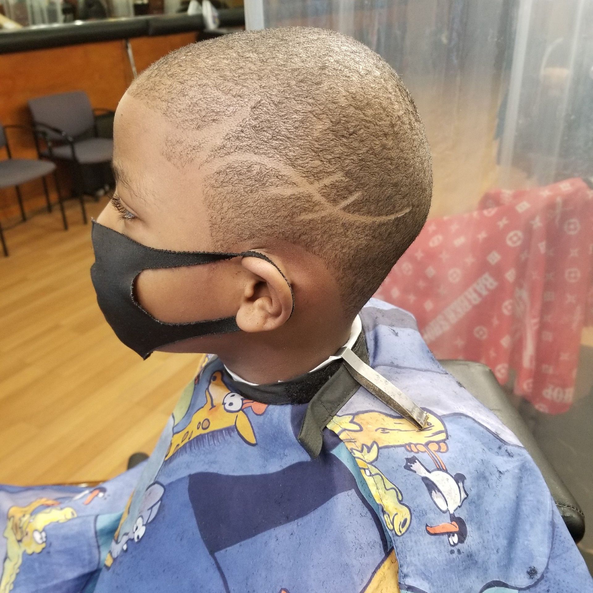 Boy with a patterned fade haircut, wearing a black mask and cape, in a barber shop.