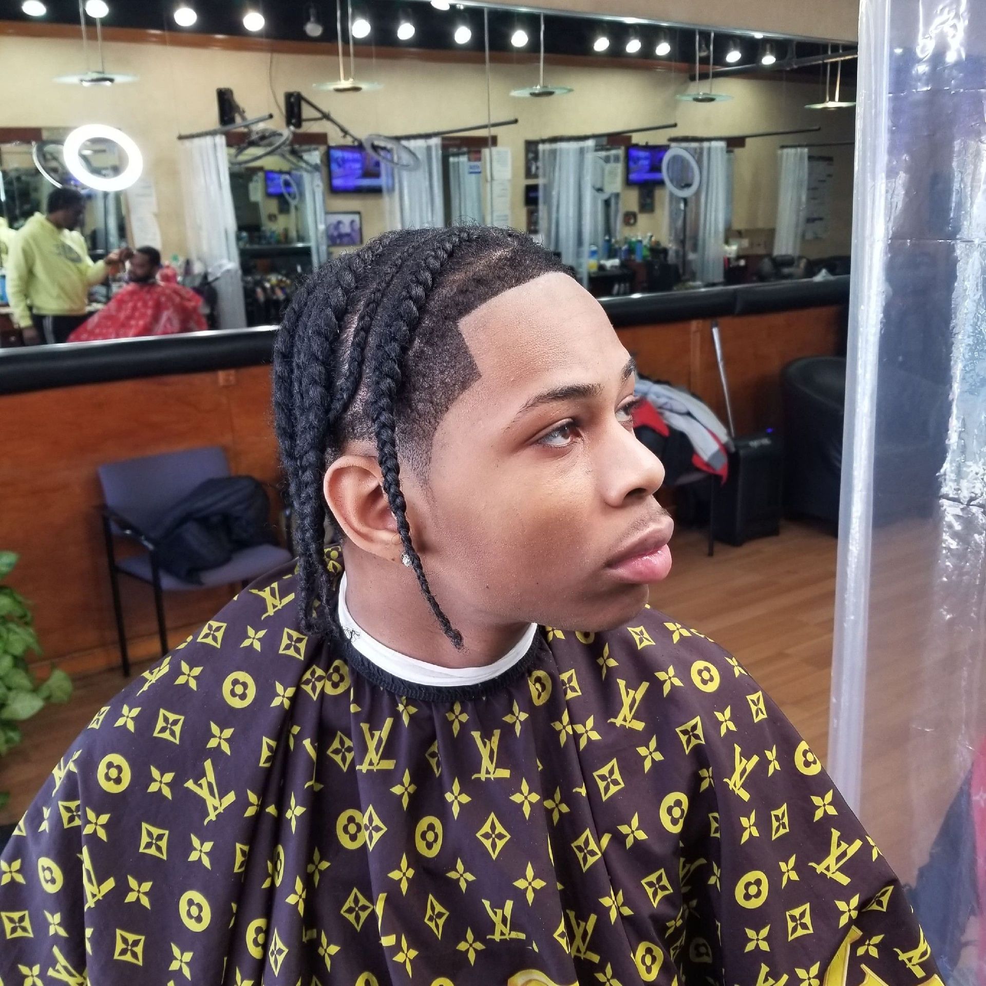 Person in a barber chair with braided hair, wearing a patterned cape. Interior barber shop with other people in the background.