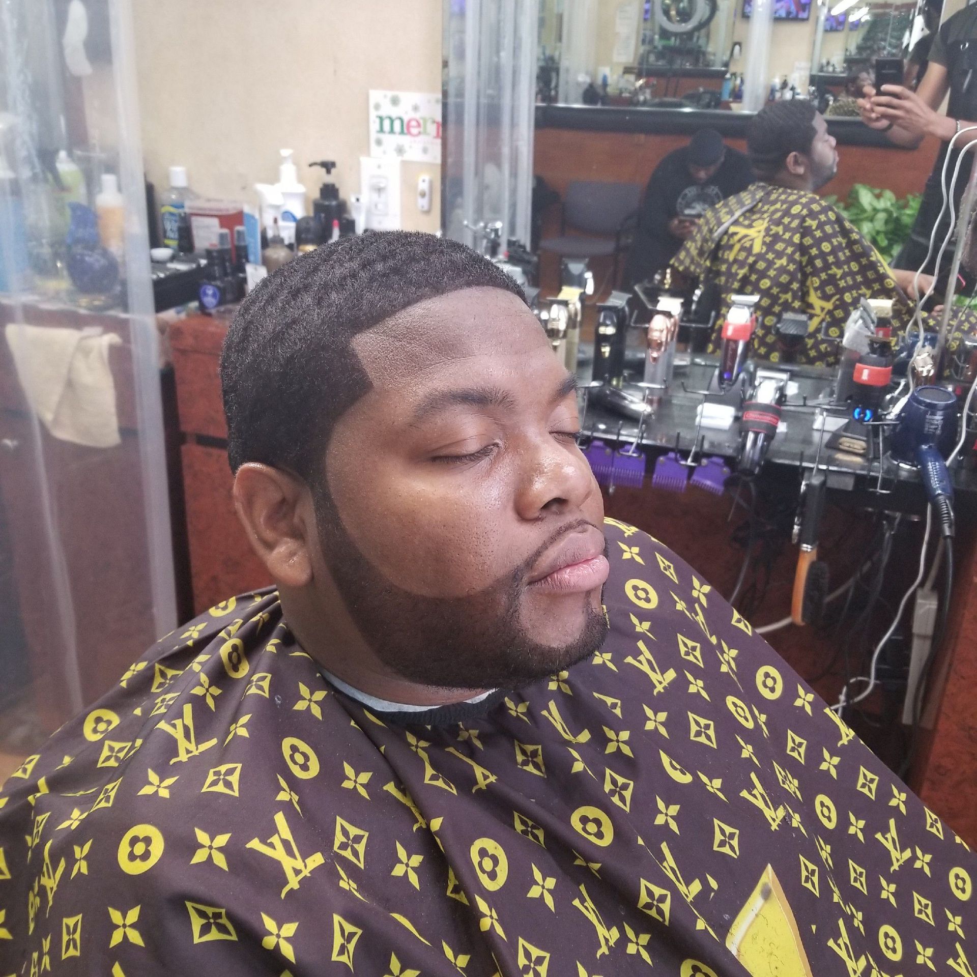 Person getting a haircut at a barber shop. He wears a Louis Vuitton cape, eyes closed.