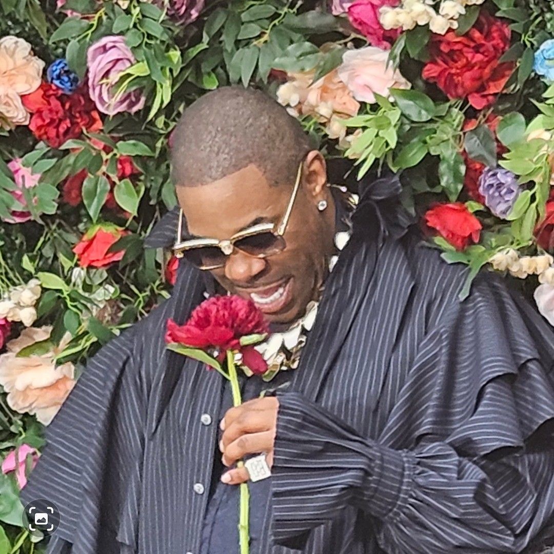 Man in sunglasses smelling a red flower in front of a flower wall, wearing a dark ruffled shirt.