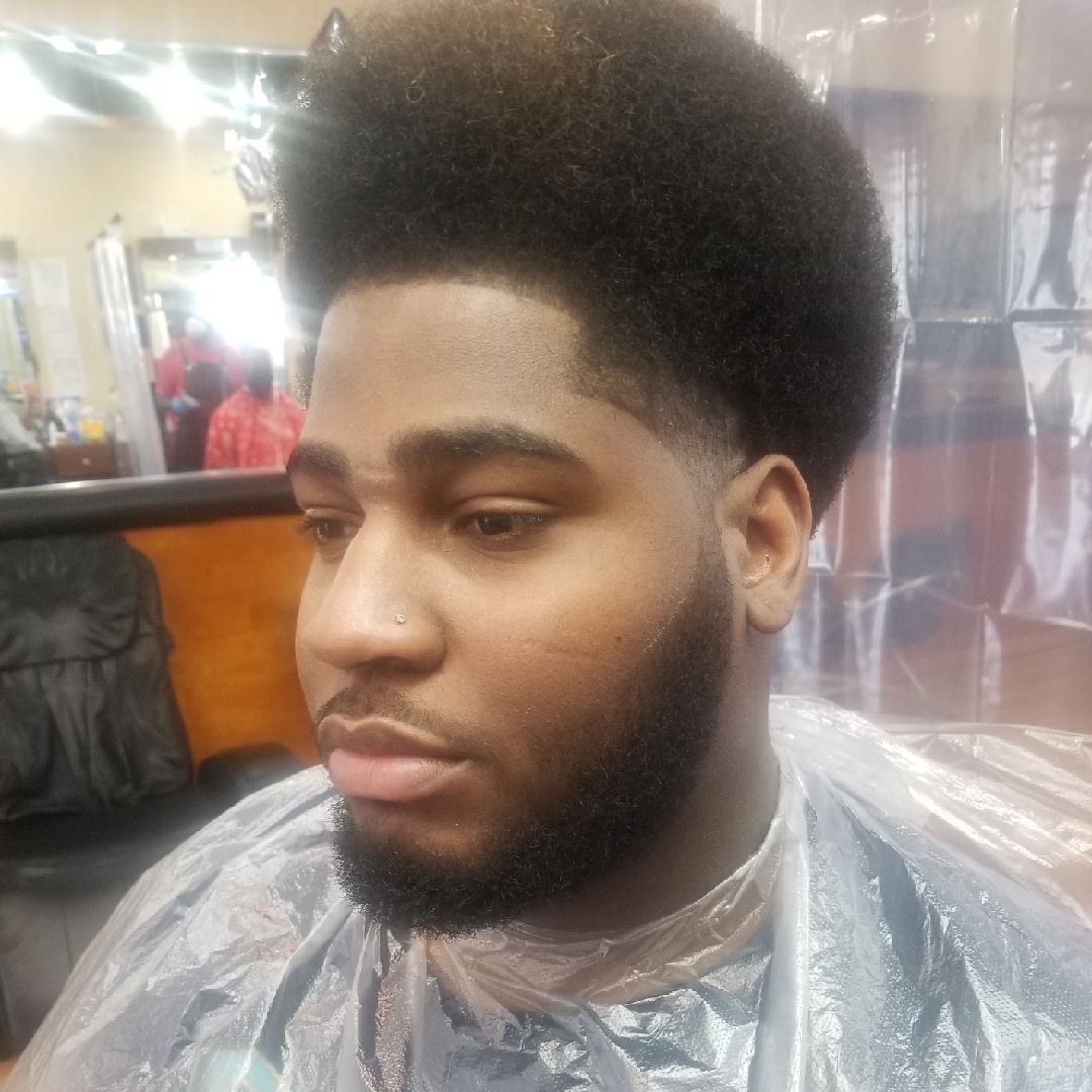 Man with a high fade haircut and beard, wearing a salon cape, looking to the side.